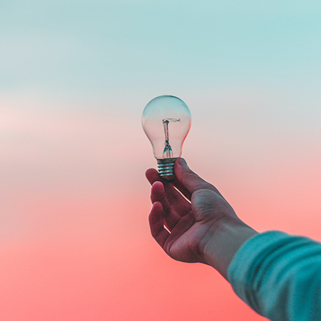 Hand som håller glödlampa mot rosa himmel. Foto från unsplash.