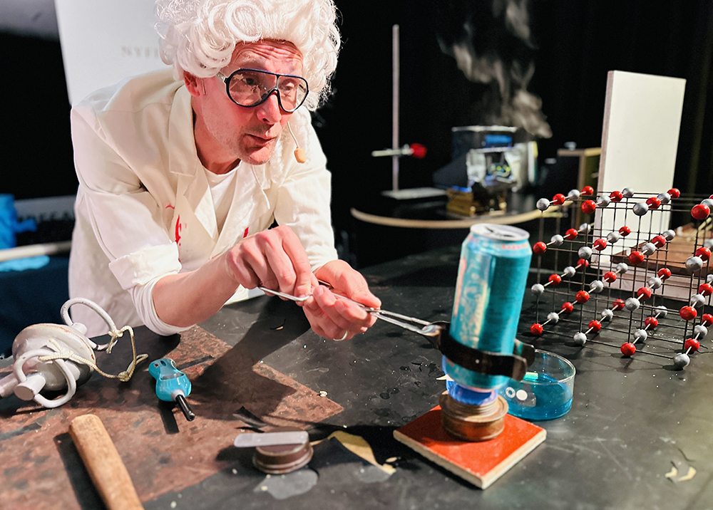 Pedagog i peruk gör kemiexperiment på Vattenhallens scen. Foto. 